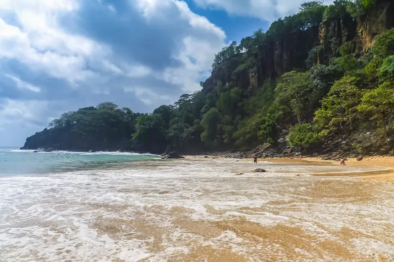 Mar avançando pela areia dourada de uma praia de Fernando de Noronha, entre falésias verdes
